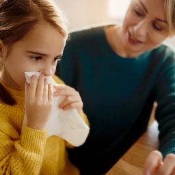 A child holding a tissue while being comforted by an adult at a table, suggesting they may not be feeling well.