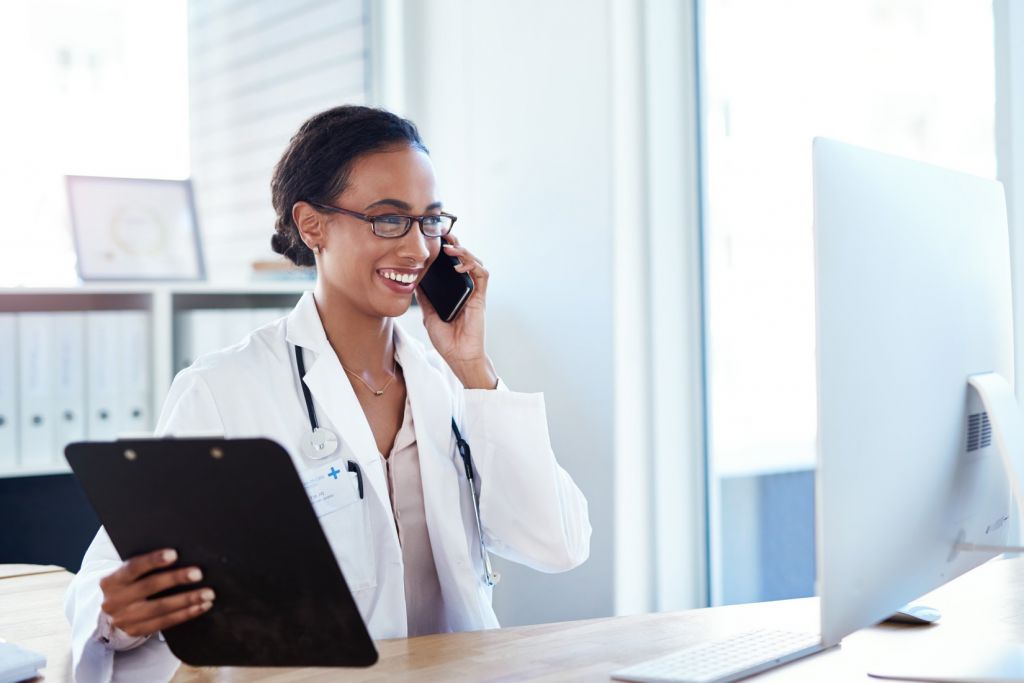 Healthcare professional speaking on the phone while holding a clipboard at a desk in a medical office.