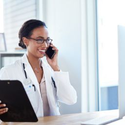 Healthcare professional speaking on the phone while holding a clipboard at a desk in a medical office.