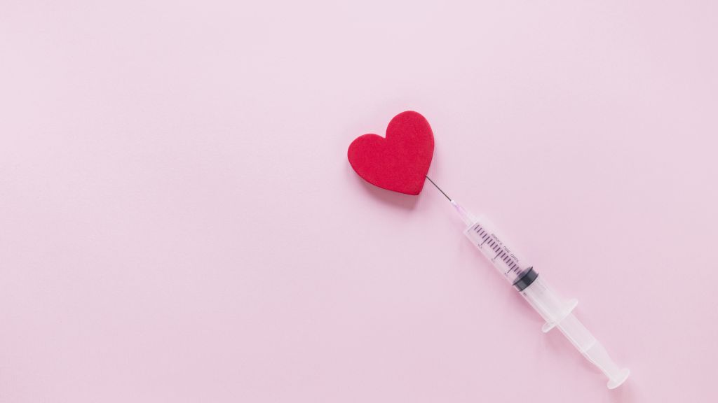 A syringe touching a small red heart on a pink background.