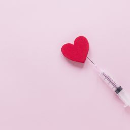 A syringe touching a small red heart on a pink background.